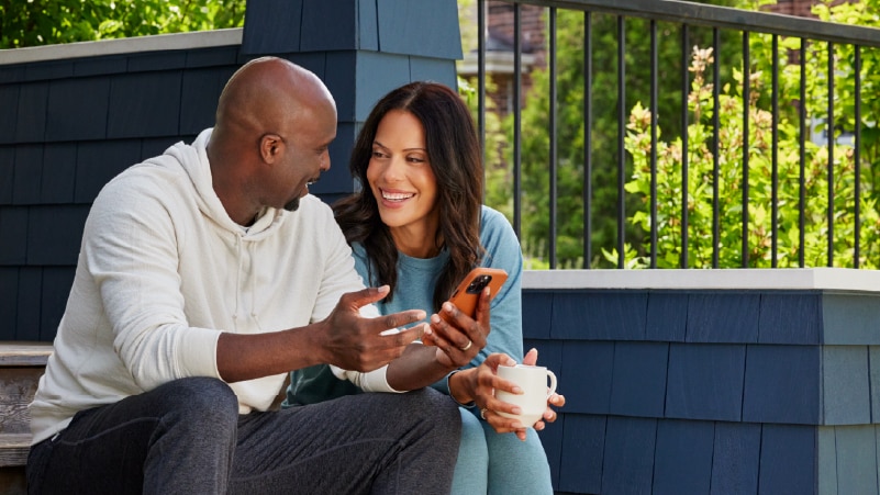Two people sit on the front steps of a house, conversing while looking at a mobile phone.