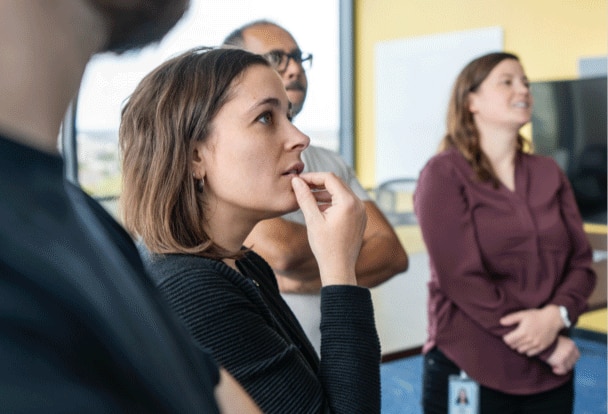 A group of business people stand together in an office looking at something in the distance.