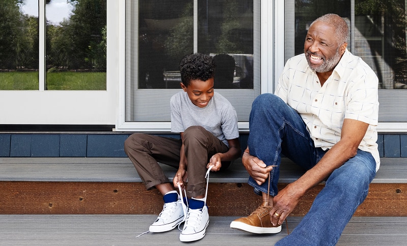 A father and son tying their shoes together, sitting in front of their home. 