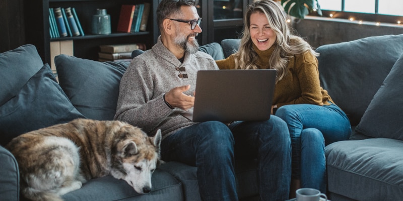 Two people sit on a couch smiling as they look at a laptop. A dog is lying next to them. 