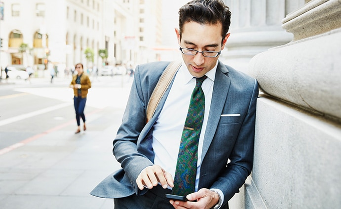 Man in business suit leaning against stone building looking at his phone