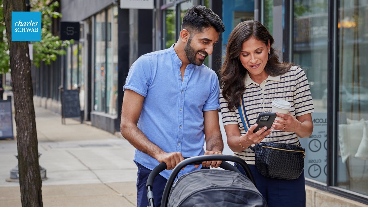 A person pushing a stroller walks beside another person holding up their phone. Both smile while looking at the phone screen.